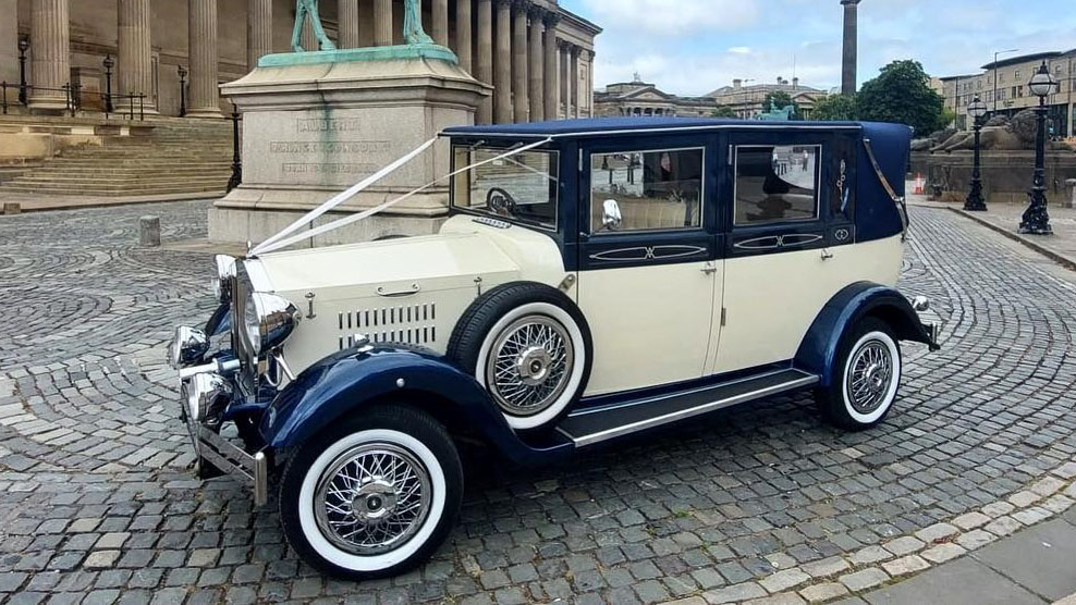 1930s Vintage Imperial in Metallic Navy Blue over Ivory dressed with white wedding ribbons across front bonnet with spare wheel mounted on the side of the vehicle parked in the street of Liverpool