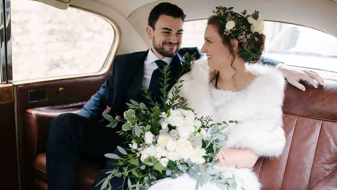 Bride wearing a white dress and holding a large bouquest of flower seating next to her Groom inside a classic Austin Princess with Brown Leather interior