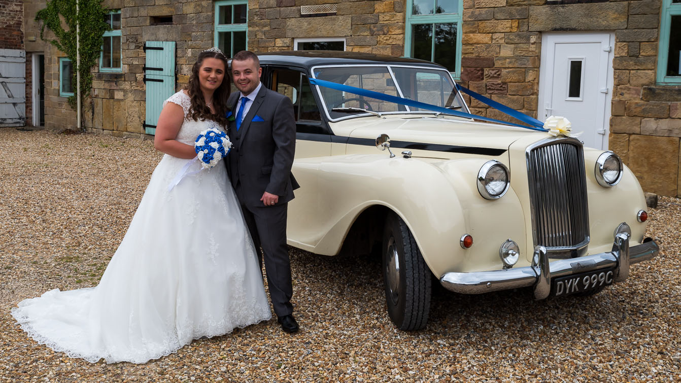 Bride and Groom standing in front of a Classic Austin Princess Princess Limousine in Ivory with Black Roof and Royal Blue Ribbons