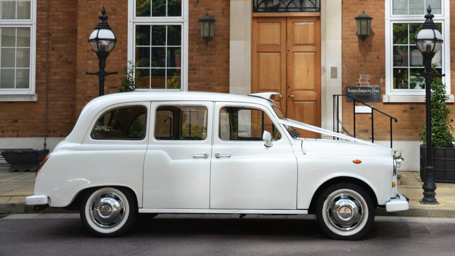 Left side view of a white classic taxi cab with white ribbons accros its bonnet and white wall tires