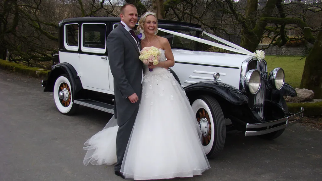 Bride and Groom standing next to the Vintage Wedding Car decorated with white ribbons