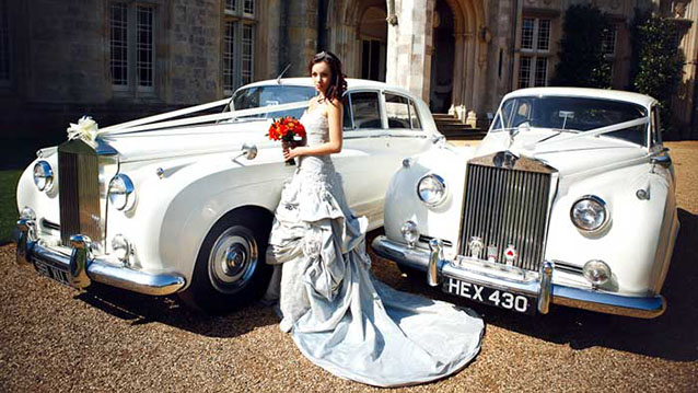 Bride in her Wedding Gown standing in front of two identical Rolls-Royce Silver Cloud decorated with White Ribbon