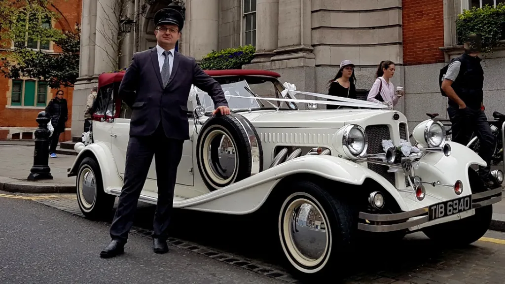 Chauffeur wearing suit, tie and chauffeurs' hat standing next to a Vintage Beauford