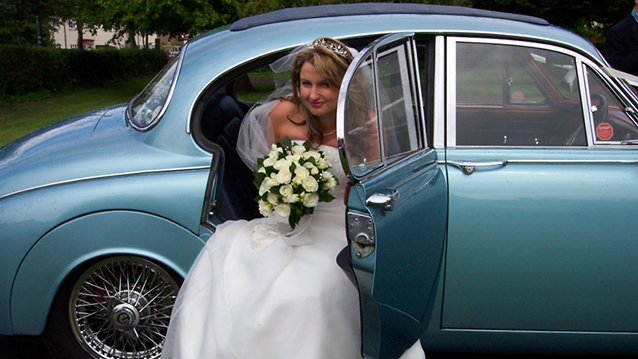Bride wearing a white dress and holding a bunch of flowers exiting the rear of the a Daimler 250 V8