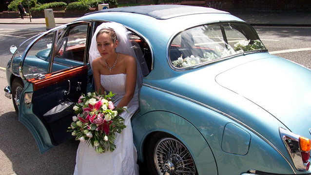 Bride wearing a white dress and holding a colourful bunch of flowers exiting the rear of the a Daimler 250 V8