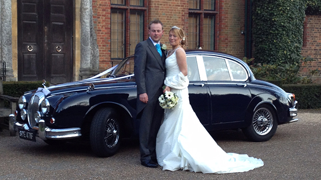 Groom holding bride in his arms with Dark blue Daimler 250 V8 in the background
