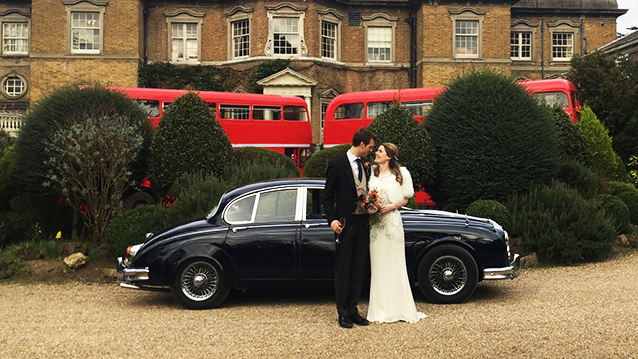 Groom holding bride in his arms with Dark blue Daimler 250 V8 in the background and a couple of Red Routemaster buses