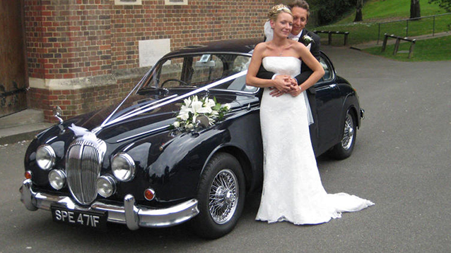 Groom holding bride in his arms with Dark blue Daimler 250 V8 in the background