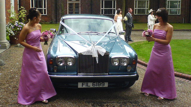 Two Bridesmaids wearing dark pink dressed in front of light blue classic Rolls-Royce Silver Shadow II