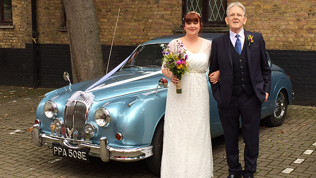 Bride and Groom standing in front of a classic blue Daimler 250 V8. Bride is holding a bridal bouquet