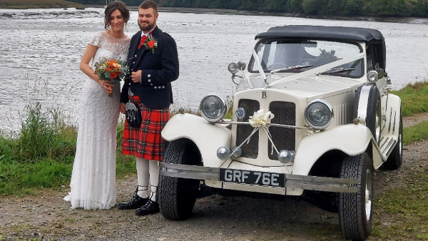 Beauford 4 Door Convertible decorated with white ribbons and black soft-top roof up. Bride and Groom standing by the vehicle. Groom wears a ceremonial kilt