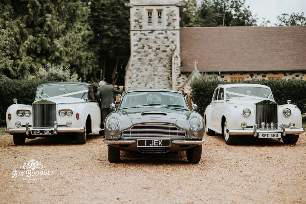Thre Classic vehicles featuring 2 classic Rolls-Royce decorated with matching white ribbons and a Classic Aston Martin DB6 in the middle. Church can be seen in the background.