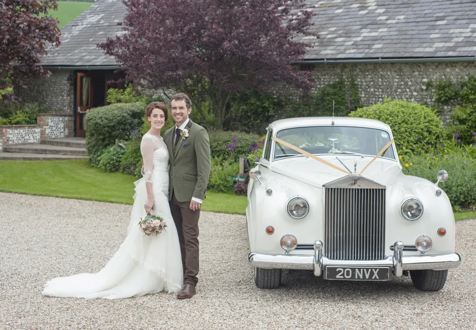 Newly Wed Couple holding each others and standing by a white classic Rolls-Royce Silver Cloud decorated with Ivory Ribbons. Upwaltham Barns entrance can be seen in the background.
