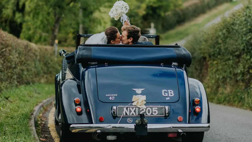 Rear view of the car with Bride and Groom inside the vehicle with the roof down kissing. Bride is waiving her bridal bouquet
