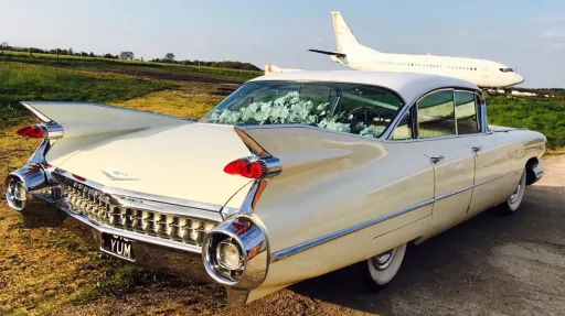 Rear view of an Ivory american Cadillac with a plane in Background. Flowers on parcel shelf can be seen trhough the rear window.