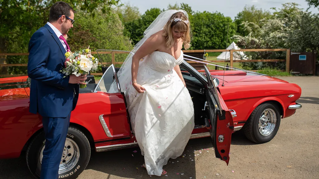 Groom holding bridal bouquet while bride comes out of the classic Ford Mustang