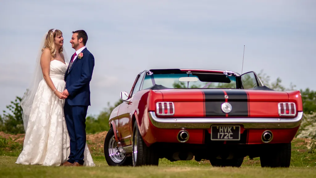 Rear view of Classic Red Ford Mustang Convertible with roof down with Brideand Groom holding hands next to the vehicle