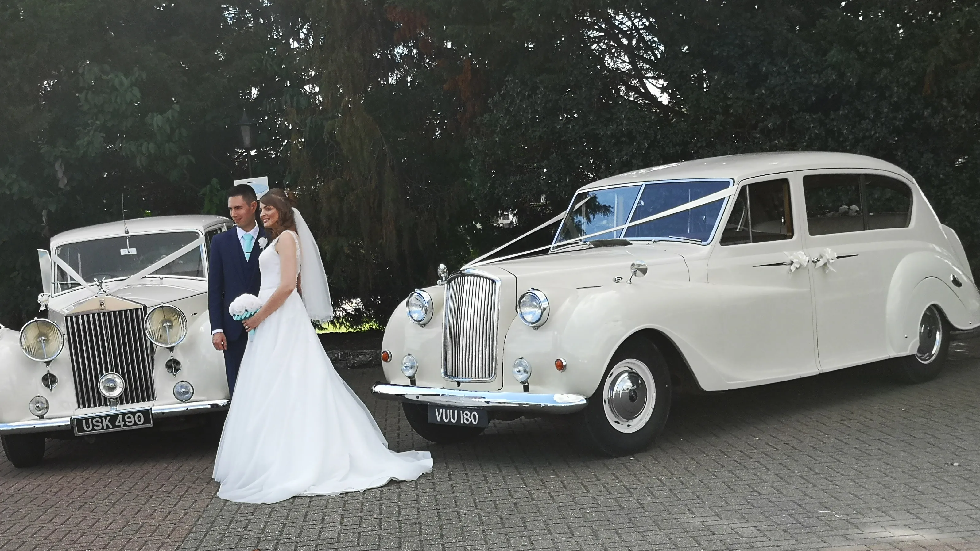 Two 7-seater Austin Princess Limousine decorated with matching ivory ribbons. Bride and Groom are stansing in the middle of the two vehicles holding each others