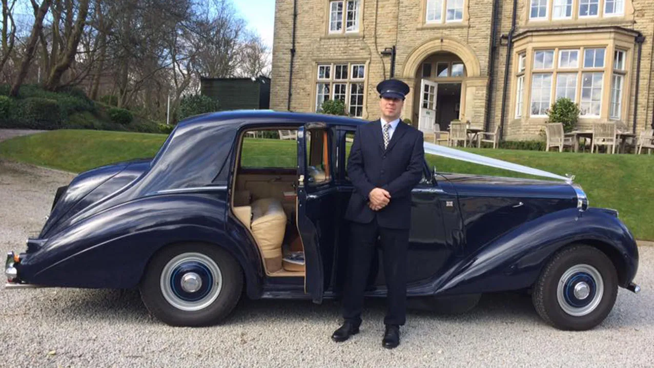 Classic Bentley R-Type in midnight blue with white ribbons and fully uniformed chauffeur standing in front of the vehicle