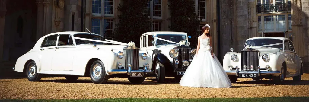 Selection of three classic and Vintage Rolls-Royce in White with Bride wearing a white wedding dress standing in the middle of the vehicles. Background is a popular wedding venue in the style of a castle in West Yorkshire.
