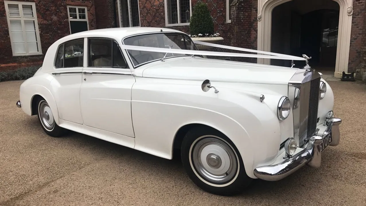 A Lincolnshire-based classic Rolls-Royce Silver Cloud in White dressed with traditional Ivory Ribbons across its front bonnet. Car has white wall tires and large chrome grill with 